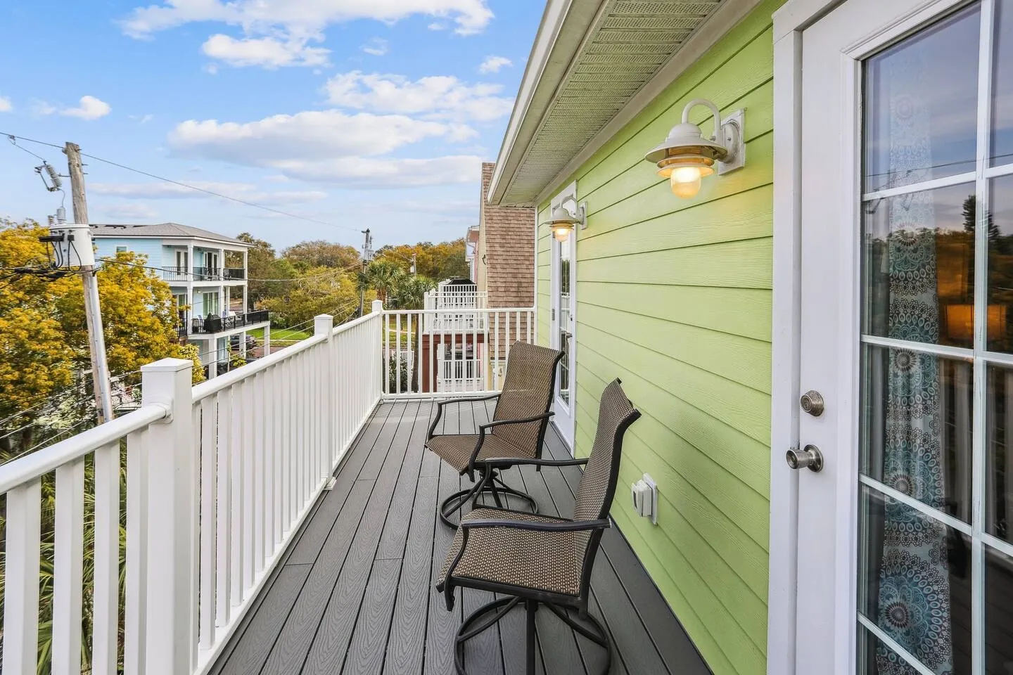 Balcony with two chairs, white railings, and light green siding overlooking nearby buildings and trees under a partly cloudy sky.