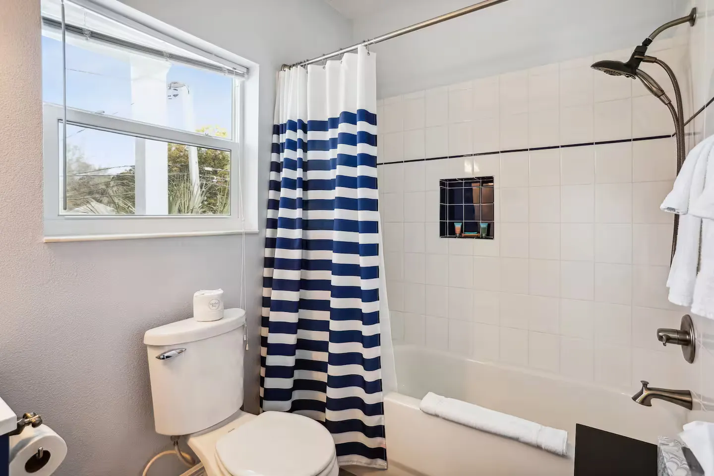 Bathroom with a toilet, window, and a bathtub shower combo featuring white tile and a blue-and-white striped curtain.