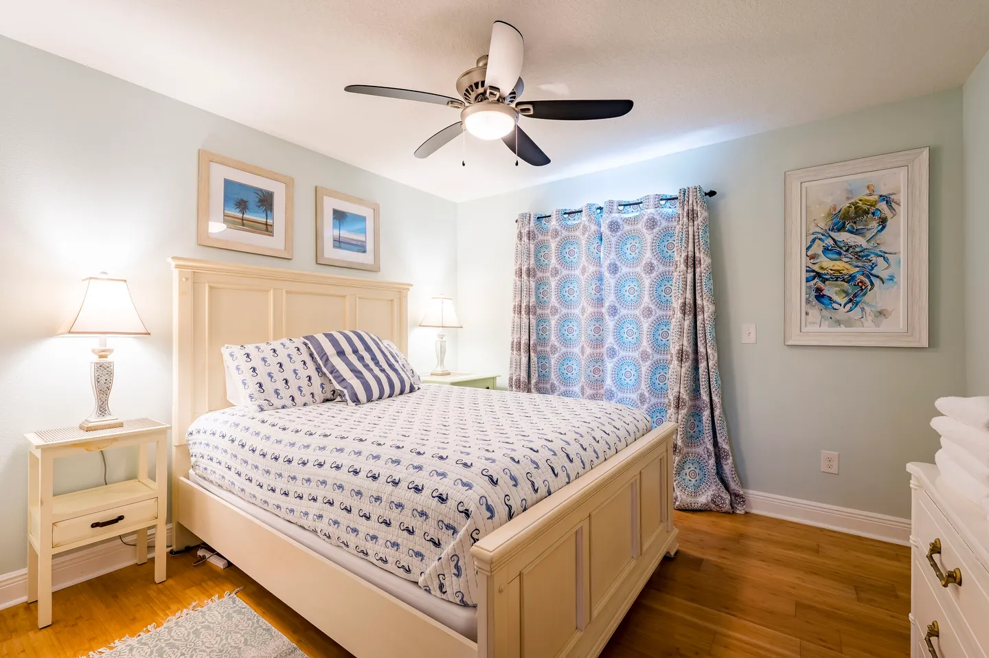 Bedroom with a white wooden bed frame, patterned bedding, matching nightstands with lamps, and a lighted ceiling fan.