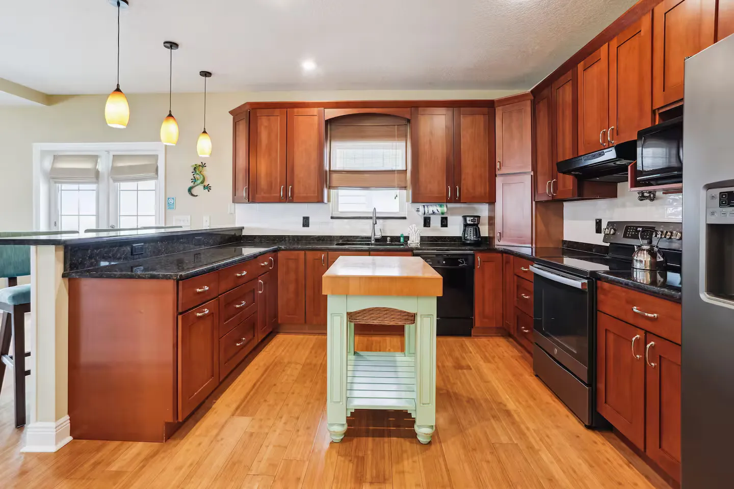 Kitchen with warm wood cabinetry, dark countertops, a light green center cart with a butcher block top, and black appliances.