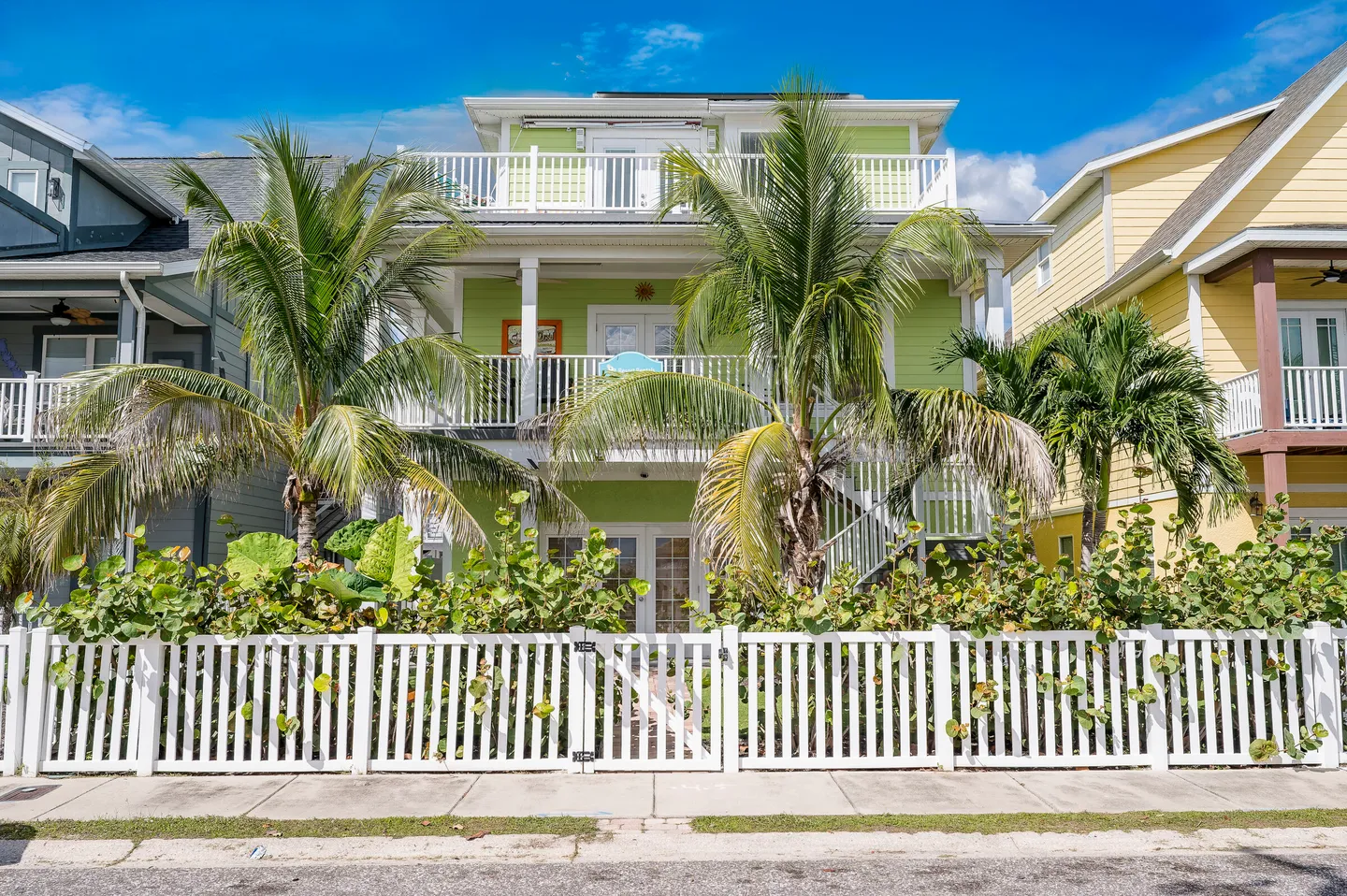 Exterior view of a three-story light green home behind a white picket fence with palm trees.