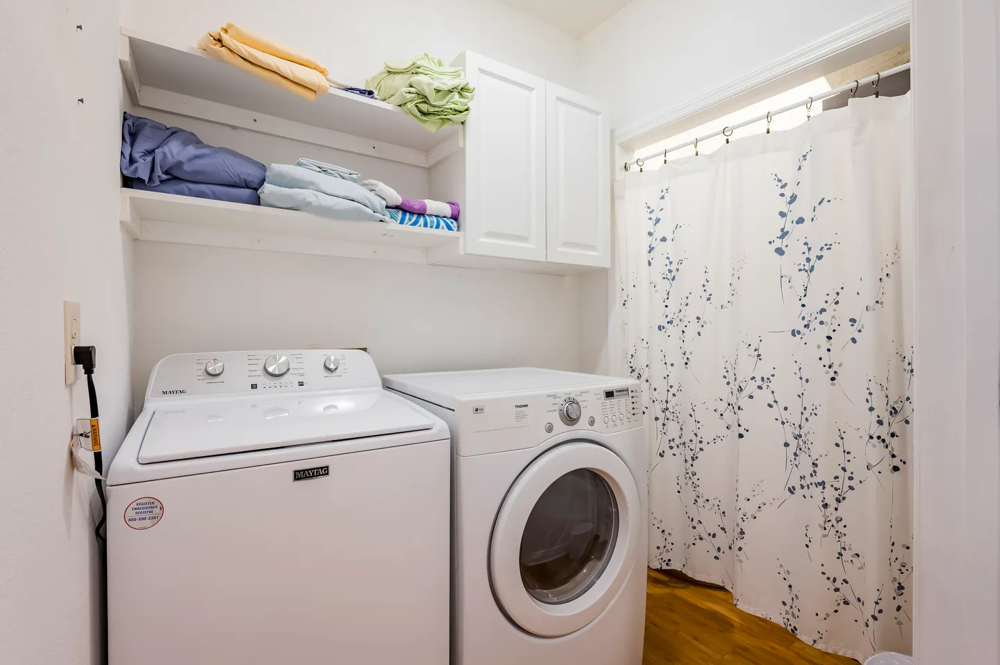 A laundry room with a white washing machine, dryer, overhead shelving with folded linens, and a curtained doorway.