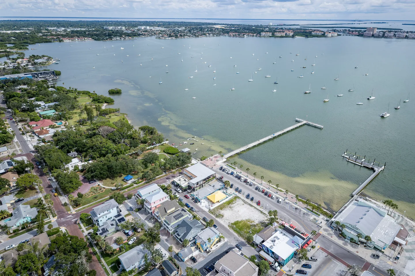 High-angle aerial view of a coastal neighborhood next to a large bay with boats moored and a long pier extending into the water.