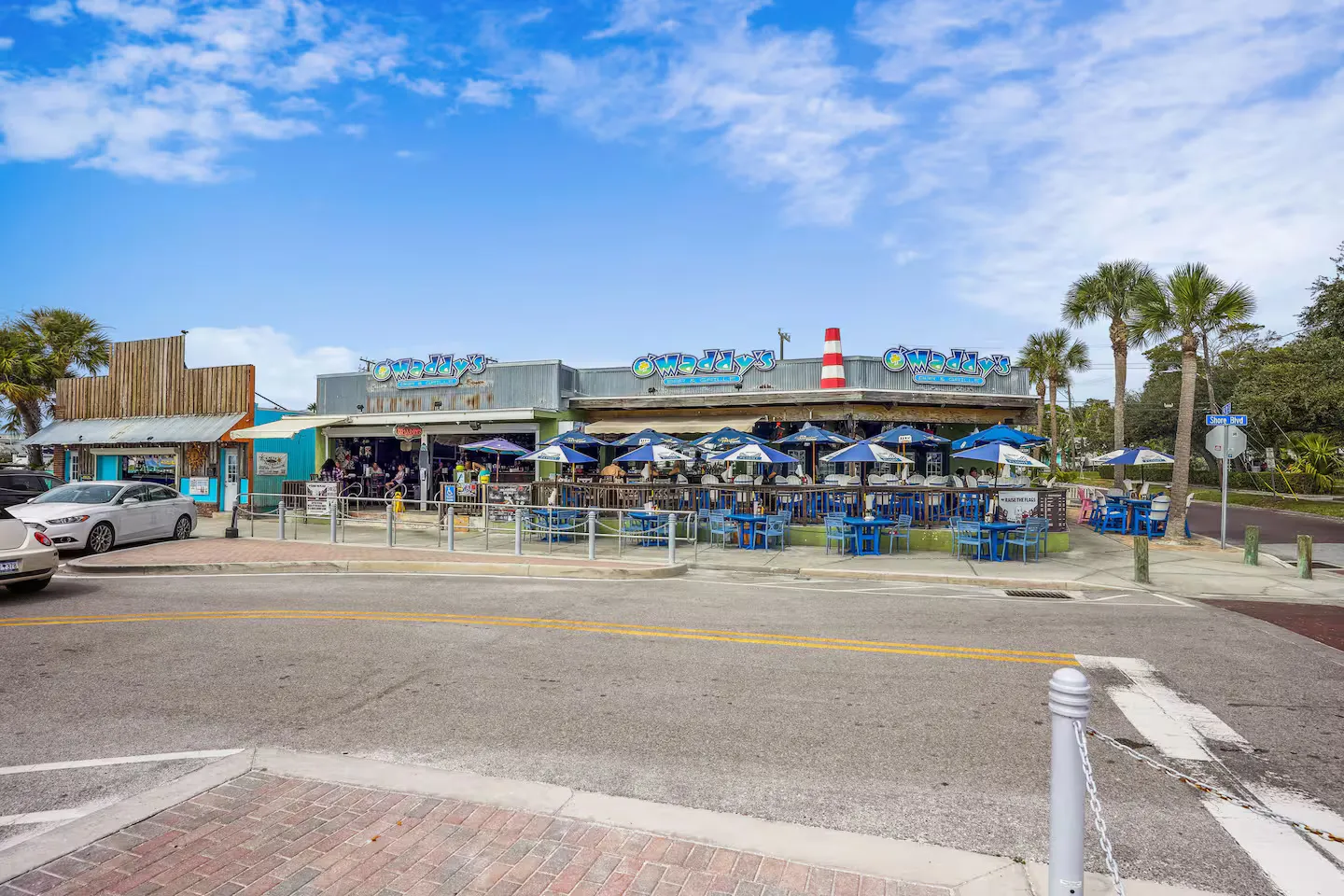 Street view of an outdoor restaurant and bar with patio seating under blue umbrellas on a sunny day.