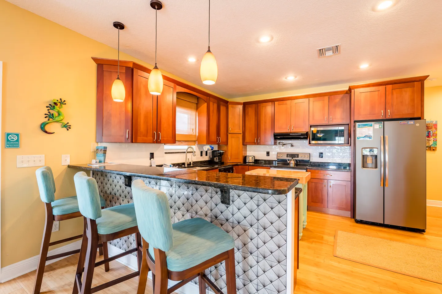 Kitchen with warm-toned wooden cabinets, dark countertops, a raised breakfast bar with three teal stools, and stainless steel appliances.