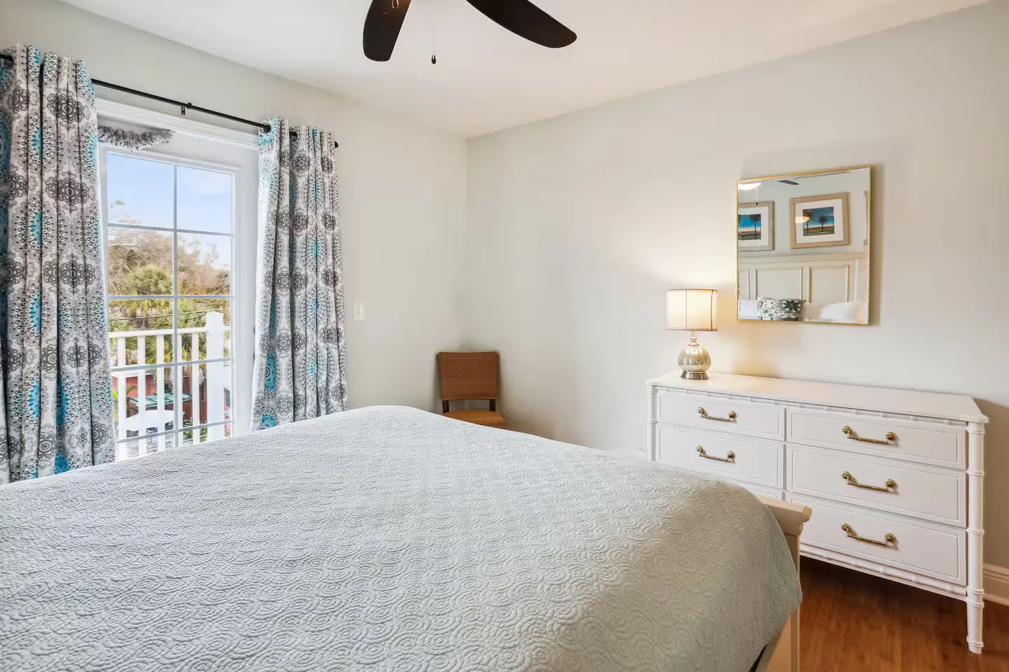 Bedroom with a patterned bedspread, white six-drawer dresser with a lamp and mirror, and a glass door leading to a balcony.
