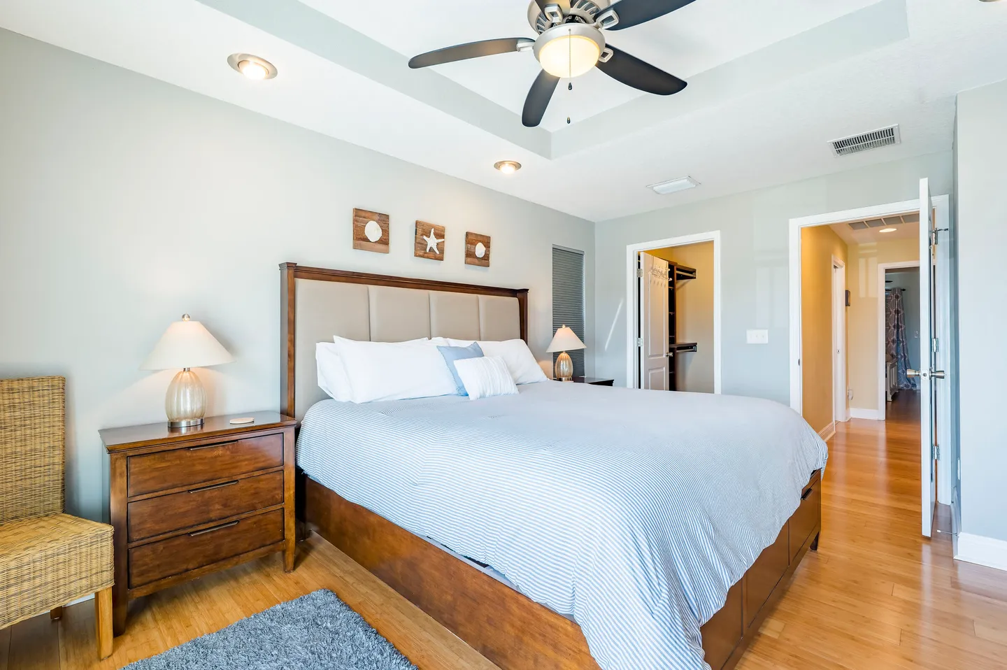 Bedroom with a large upholstered bed, wood bedside table with lamp, tray ceiling with fan, and closet doorway.