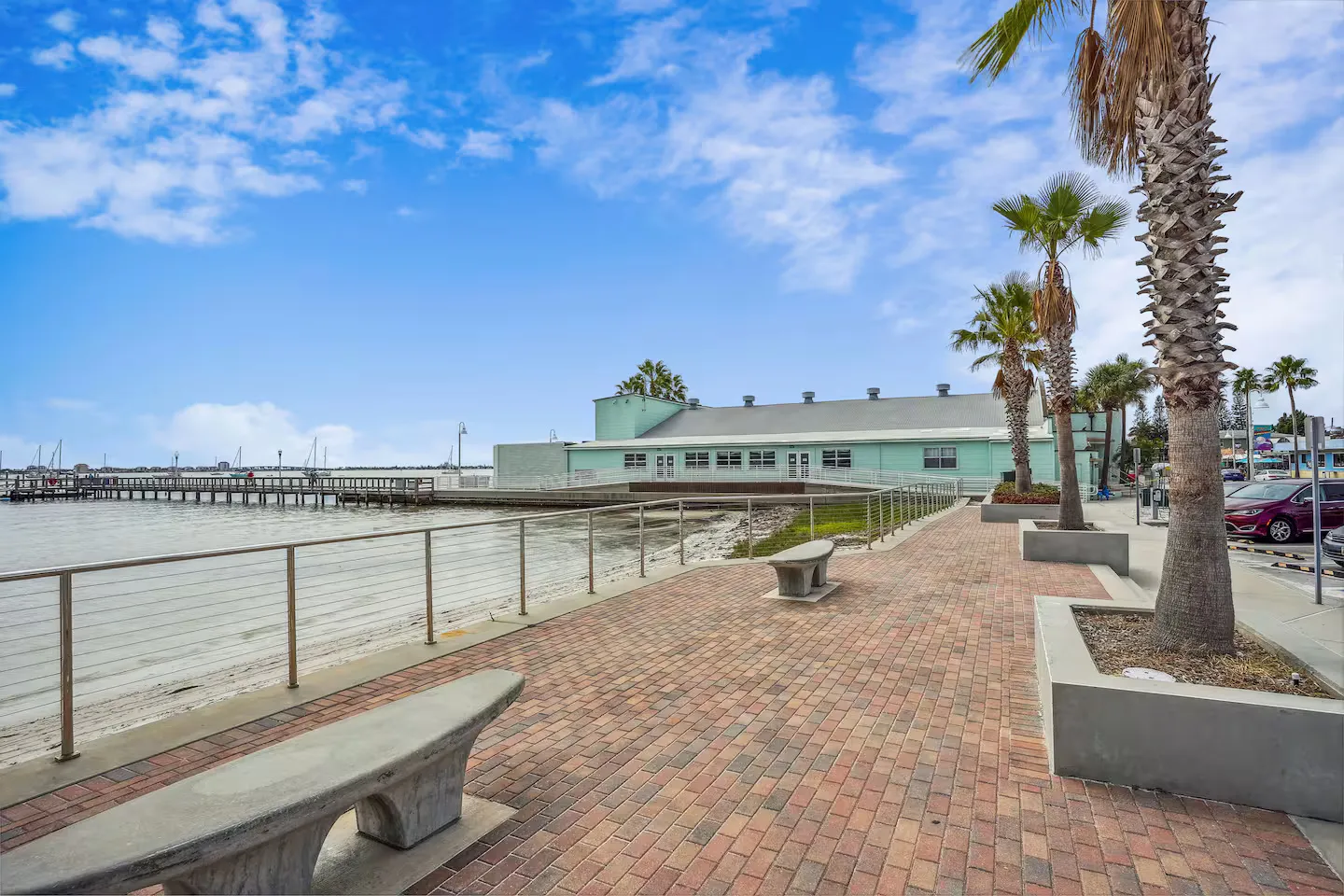 A paved waterfront promenade with concrete benches, palm trees, and a view of a light green coastal building and a wooden pier.