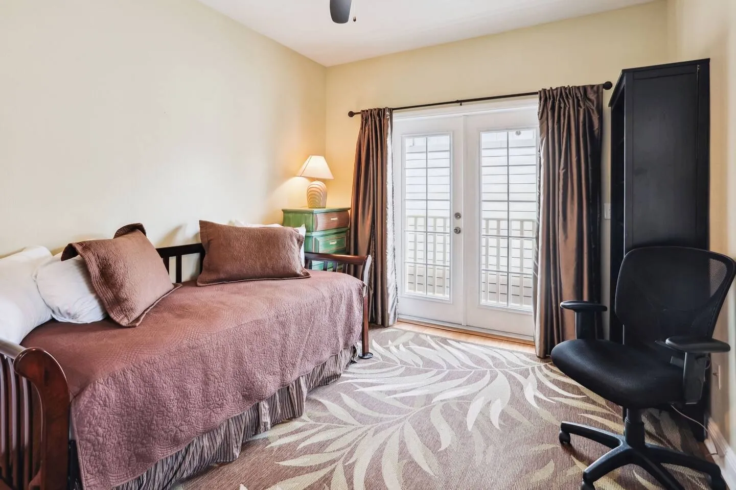 Bedroom featuring a daybed with brown bedding, an office chair, a patterned rug, and French doors leading to a balcony.