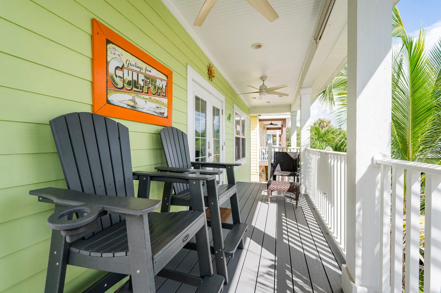 A covered balcony with lime green siding, Adirondack chairs, ceiling fans, and a white railing overlooking palm trees.