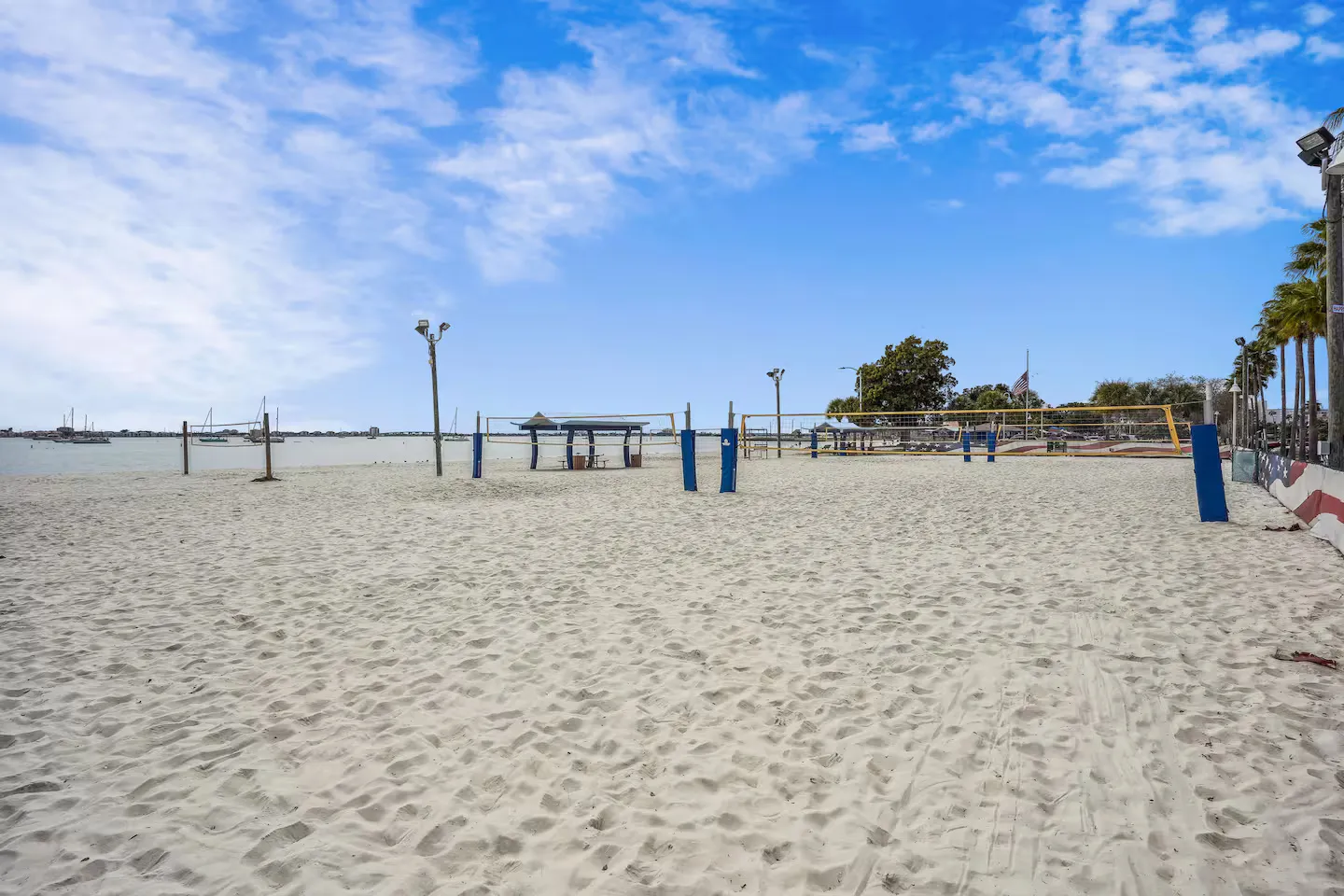 A wide sandy beach featuring sand volleyball courts with nets set up, overlooking calm coastal water with anchored boats under a blue sky.