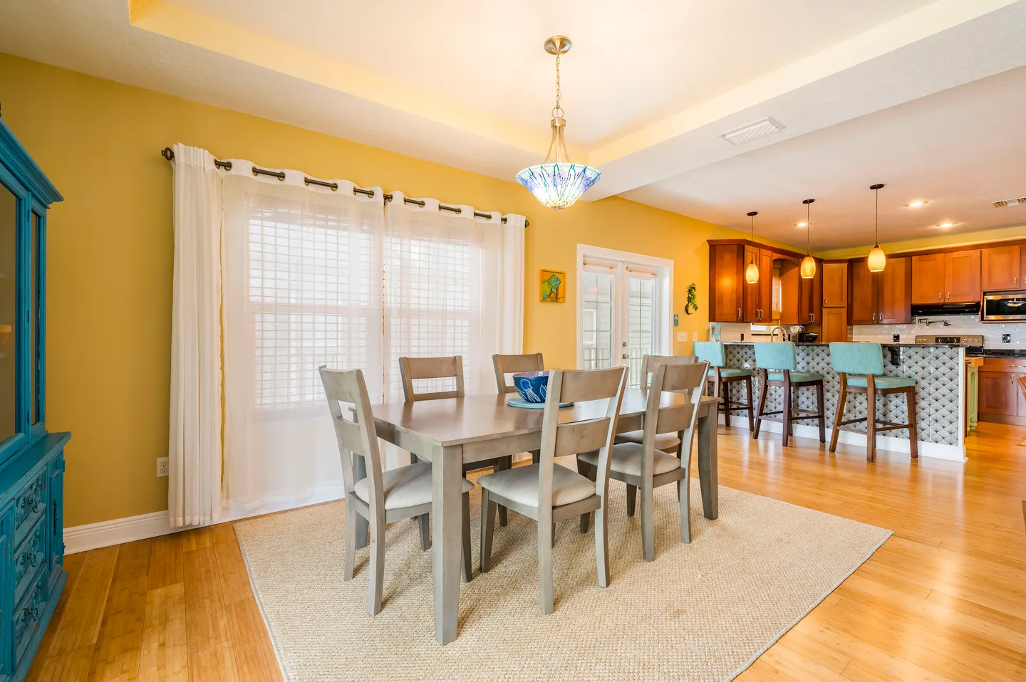 Dining area with a wooden table and six chairs placed on an area rug, adjacent to an open kitchen with breakfast bar seating.