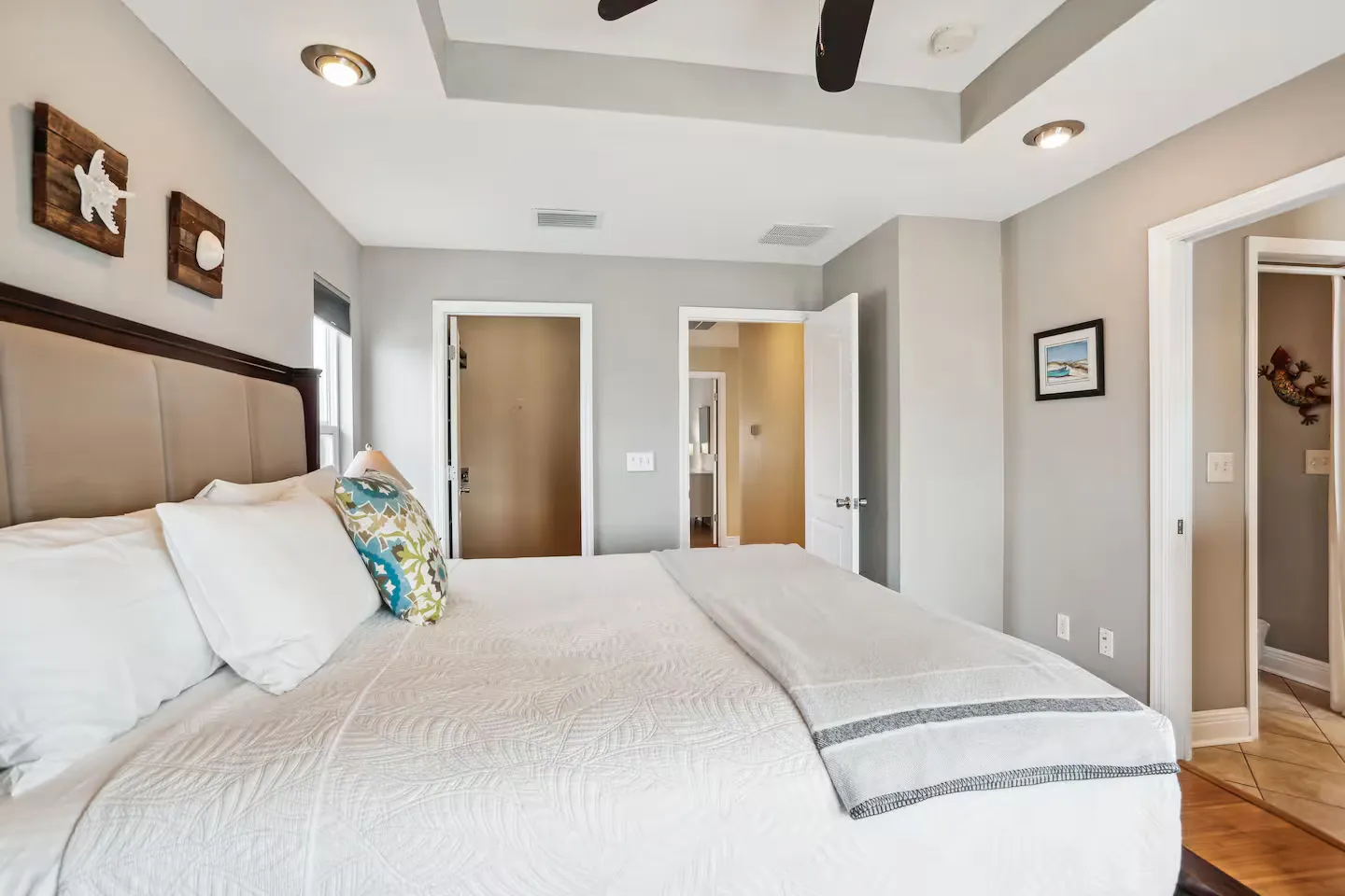 Bedroom with a neatly made bed, recessed tray ceiling, and open doorways leading to an adjoining bathroom and hallway.