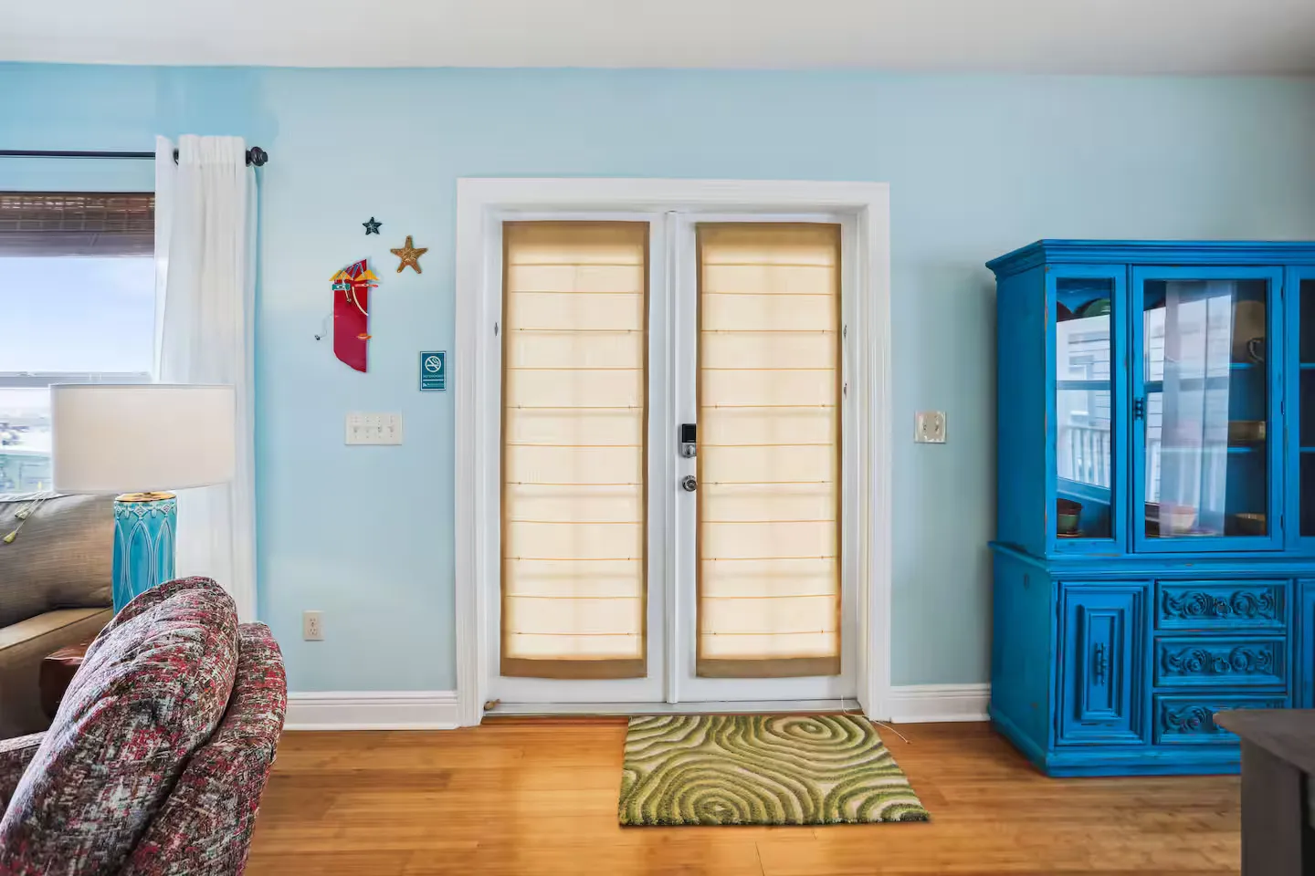 Living room entryway with light blue walls, double glass doors fitted with shades, a vibrant blue hutch, and wood-look flooring.