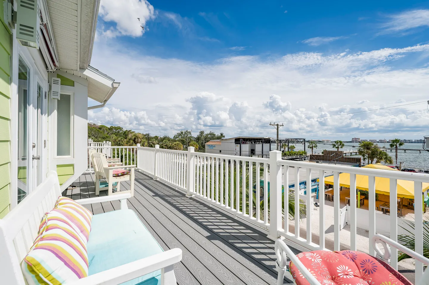 Outdoor balcony with grey decking, white railings, cushioned seating, and a view of the surrounding area and water.