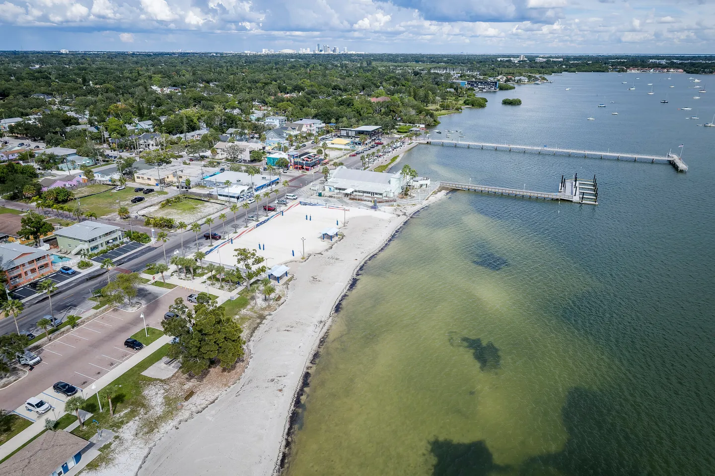 Aerial view of a coastal beach area with volleyball courts, piers extending into the water, and surrounding neighborhood.