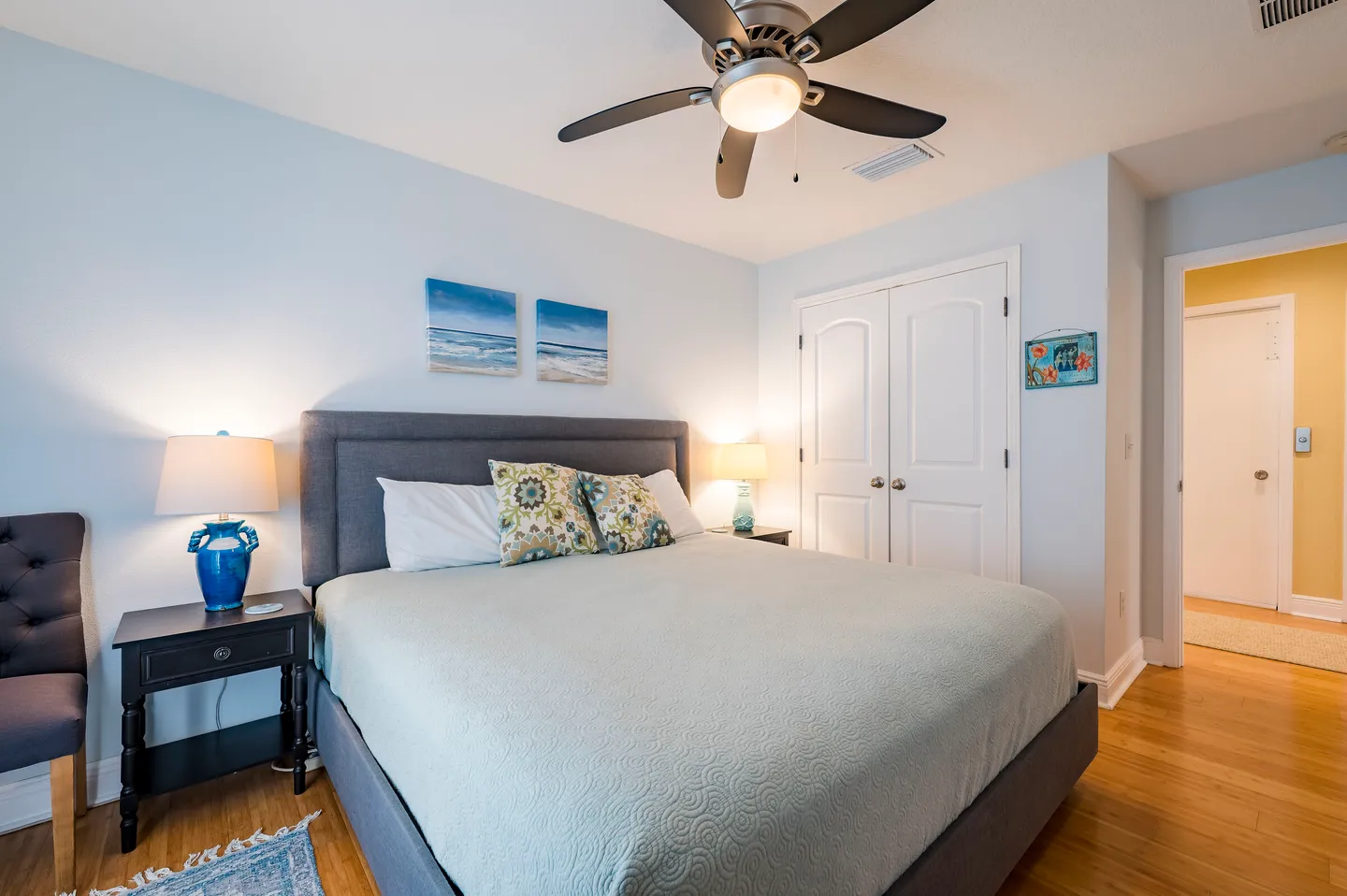 Bedroom with an upholstered gray bed, ceiling fan, bedside tables with lamps, and closet doors.
