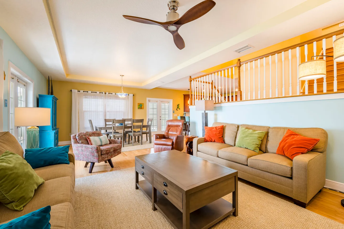 Open-concept living room with beige sofas, wooden coffee table, dining area in the background, and half-wall with railings leading upstairs.