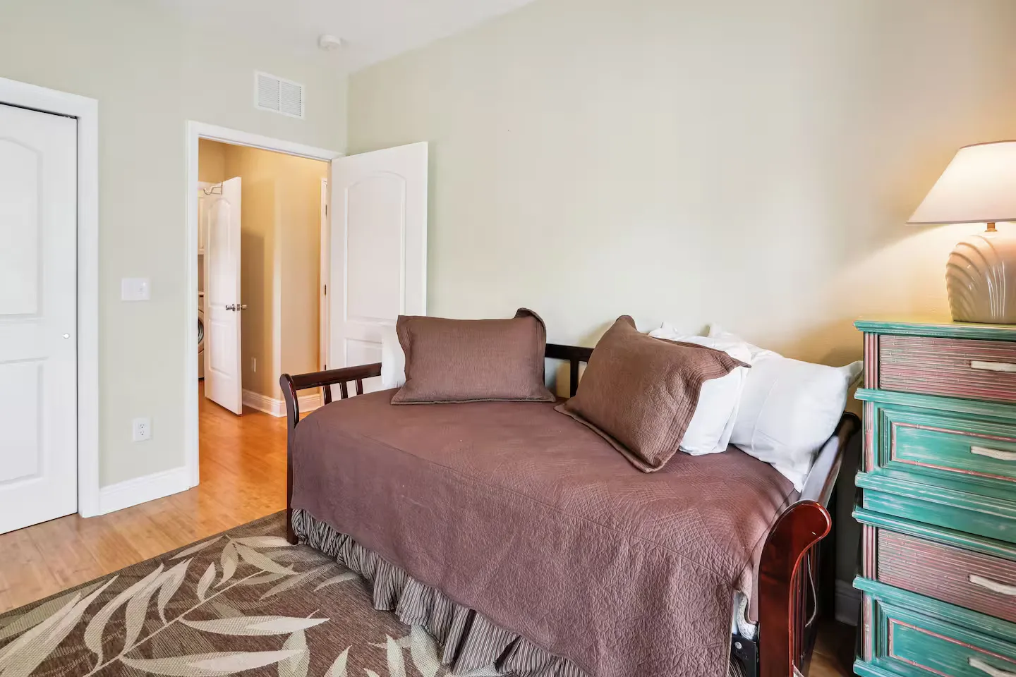 Bedroom featuring a daybed with brown bedding, an open doorway to a hallway, and a turquoise chest of drawers with a lamp.