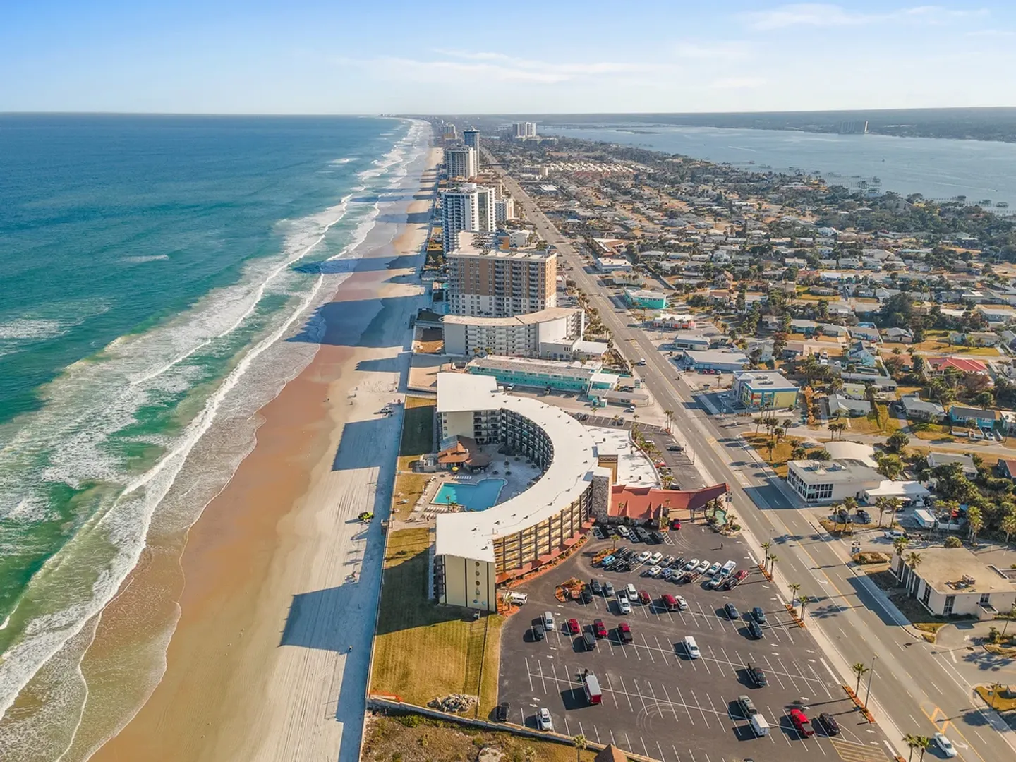 Aerial view of a curved oceanfront resort building along a sandy beach and coastline.