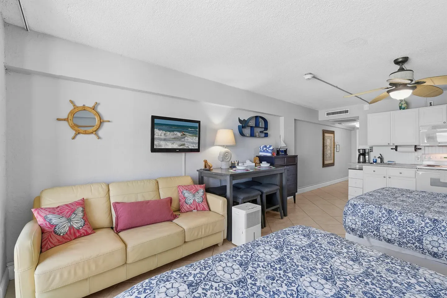 Studio living area featuring a yellow sofa, small dining table, beds with blue patterned quilts, and kitchen area.