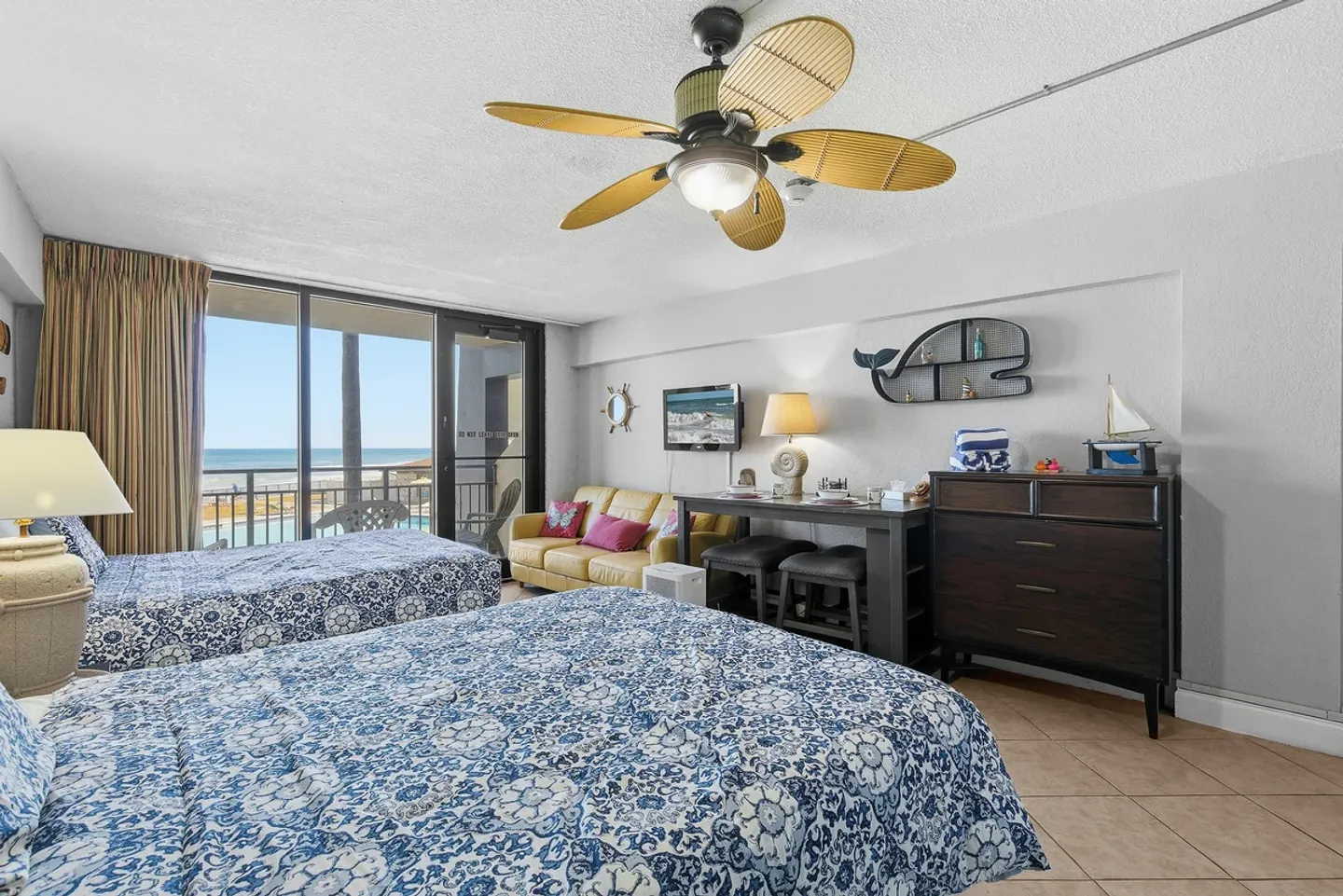 Bedroom with two beds with blue and white patterned bedding, a ceiling fan, a yellow sofa, and sliding glass doors leading to a balcony with an ocean view.