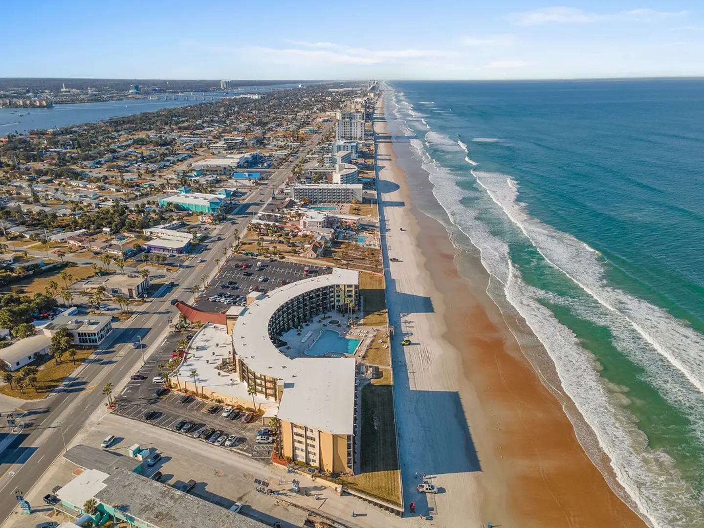 High-altitude aerial view of an oceanfront curved resort building along a sandy beach and ocean waves.