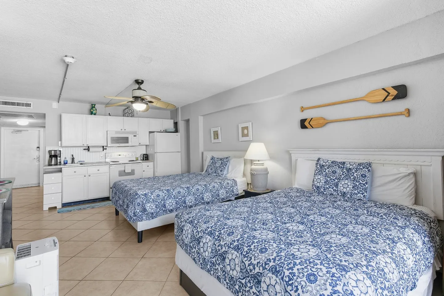 Studio rental with two queen-style beds with blue and white patterned covers, tiled floors, and a kitchenette along the wall.