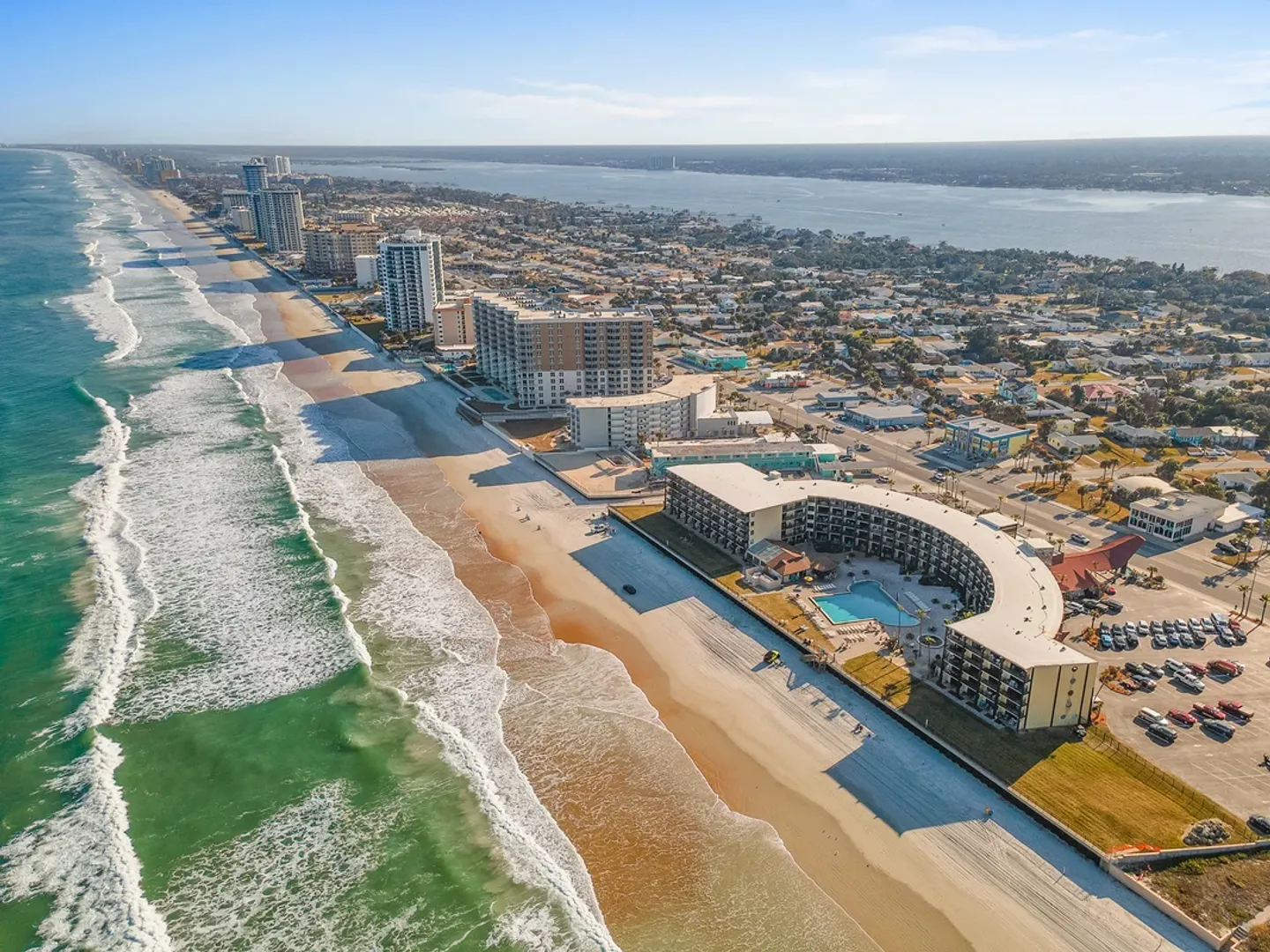 Aerial view of a coastal beach lined with oceanfront resorts, a sandy shoreline, and crashing ocean waves under a blue sky.