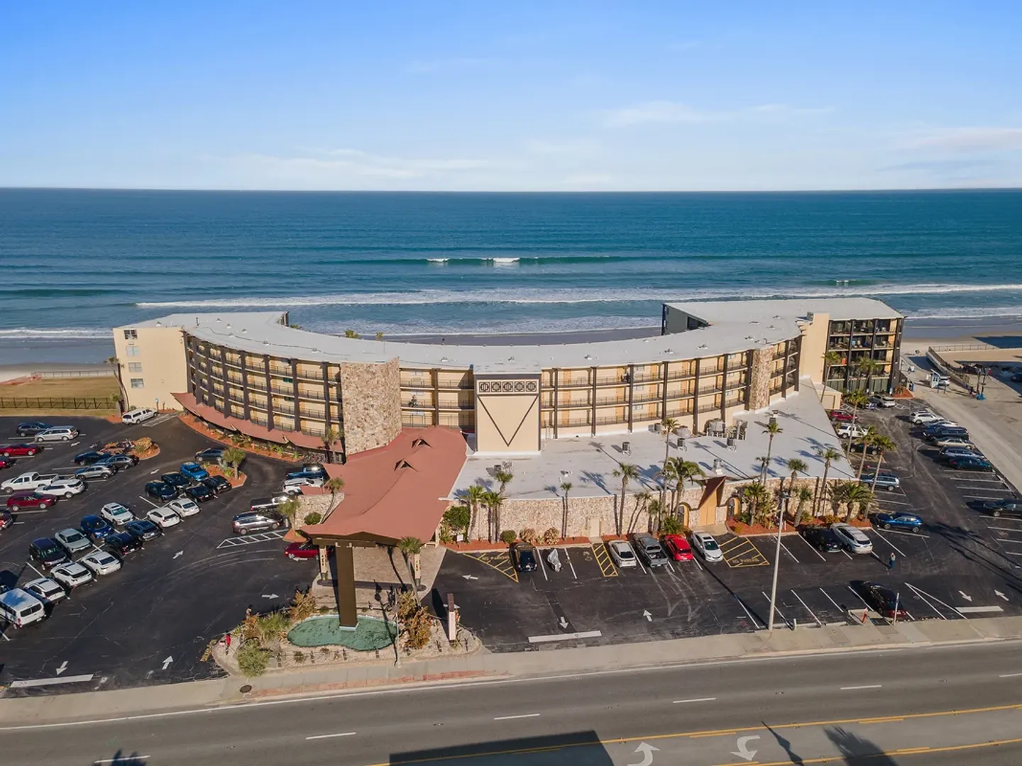 Aerial view of a curved oceanfront building with parking lots in front and the beach and ocean directly behind it.