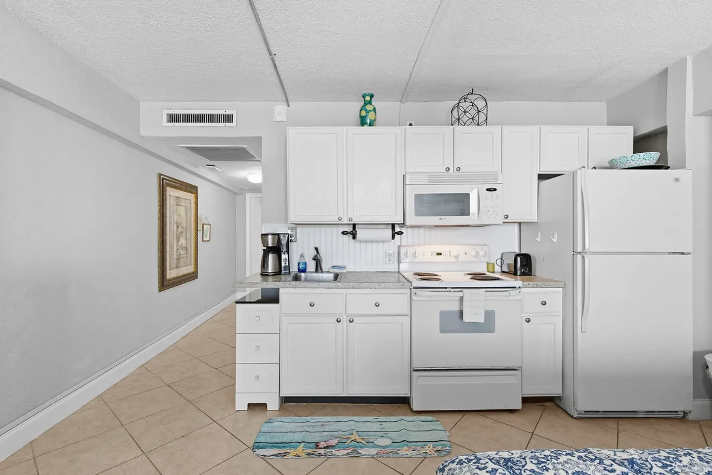 Compact kitchen area with white cabinetry, white appliances, and a tiled floor opening into a hallway.