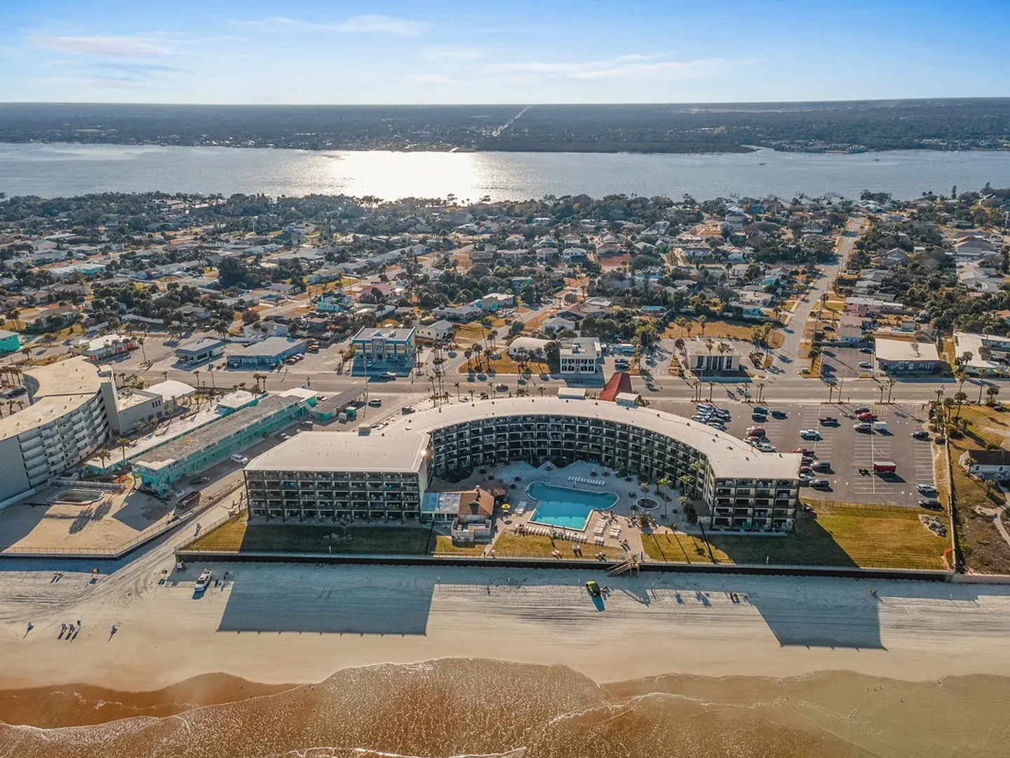 Aerial view of a curved beachfront condo complex with a swimming pool facing the sandy beach and ocean, overlooking the surrounding coastal community and inlet.