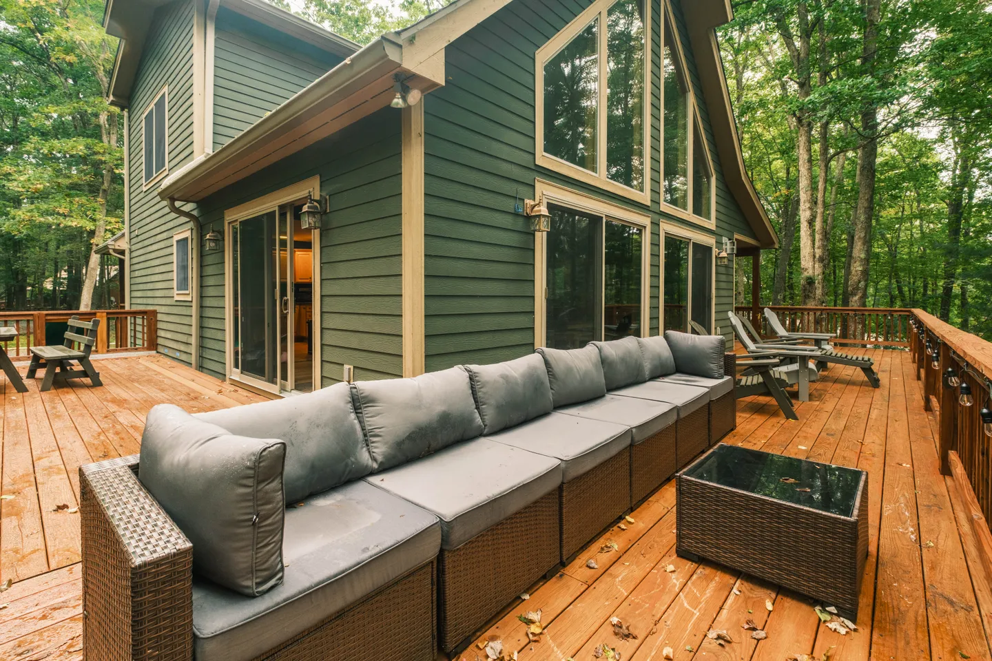 Spacious wooden back deck of a green-sided house featuring an outdoor sectional sofa and surrounded by trees.