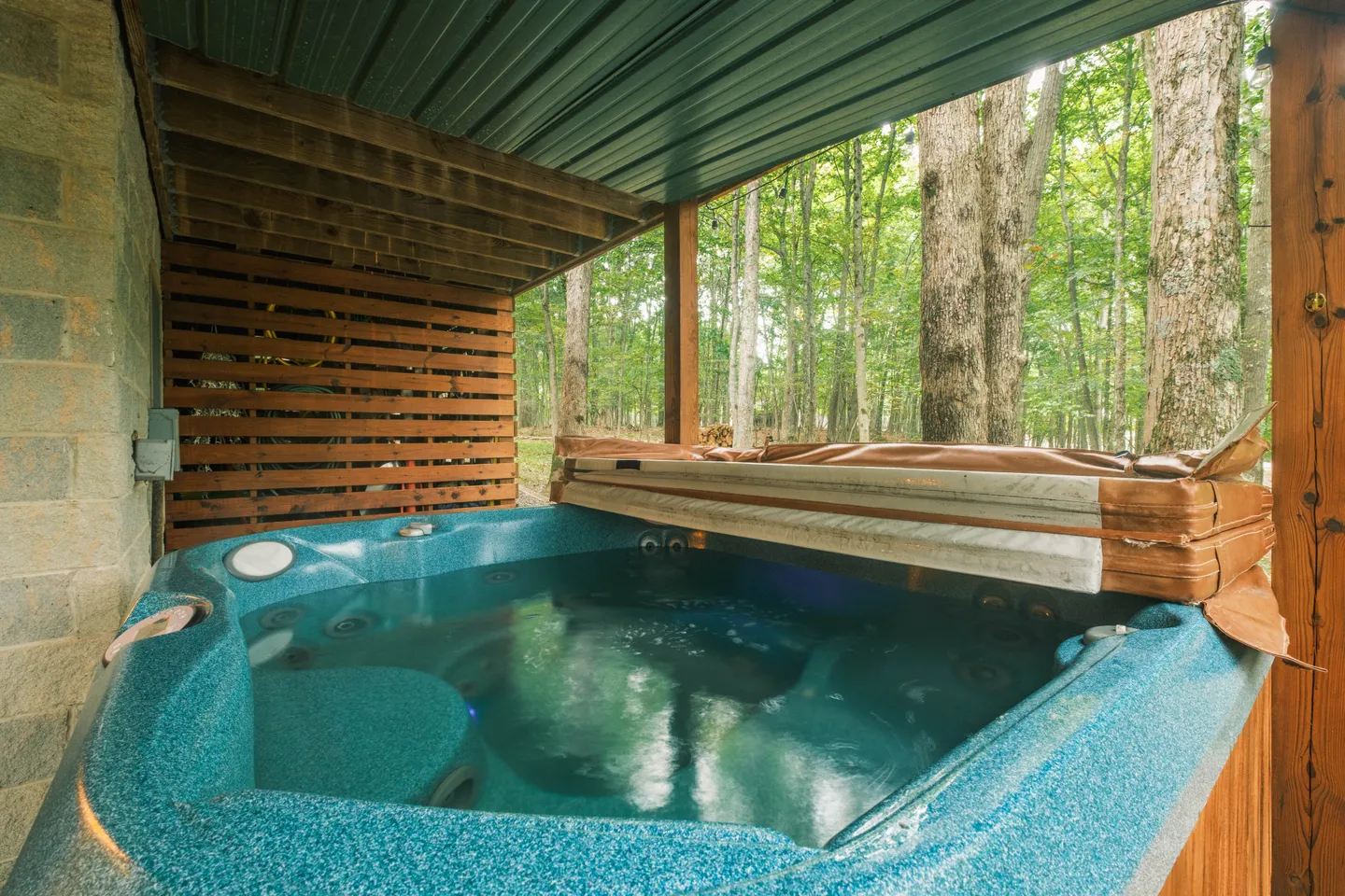 Outdoor hot tub filled with water located beneath a covered deck with a view of a wooded forest.