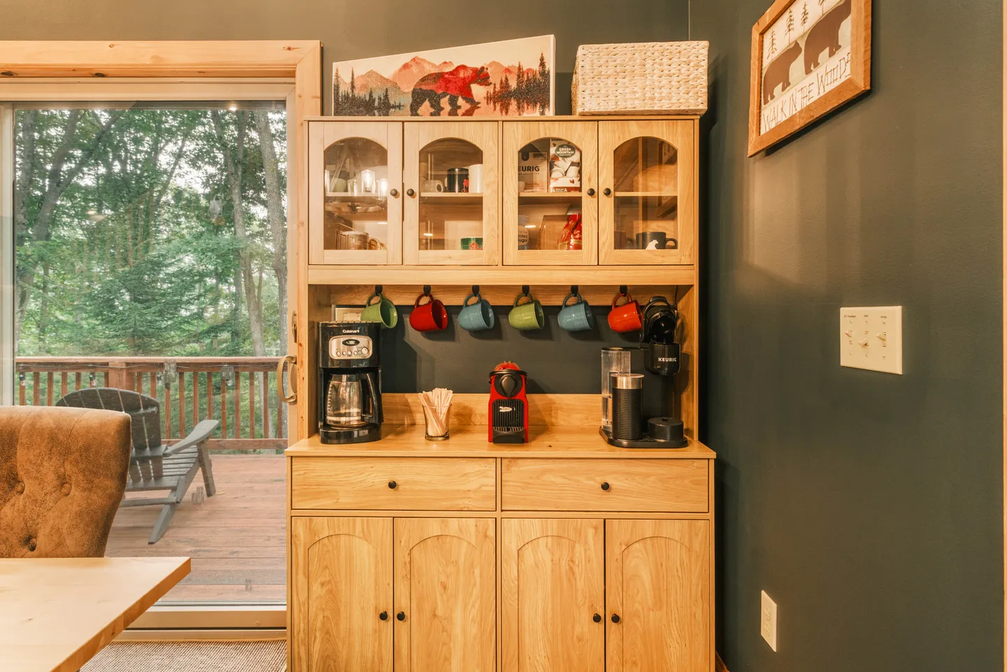 Light wood hutch set up as a coffee station with multiple coffee makers, mugs on hooks, and glass-front upper cabinets next to a sliding glass door.