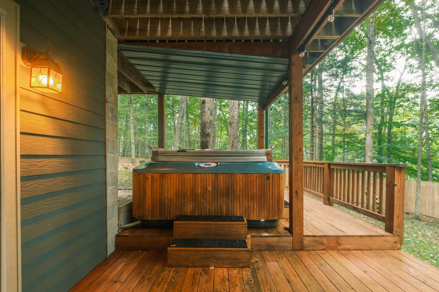 A covered outdoor wooden deck with a wood-paneled hot tub nestled beneath an upper deck, overlooking a forest of trees.