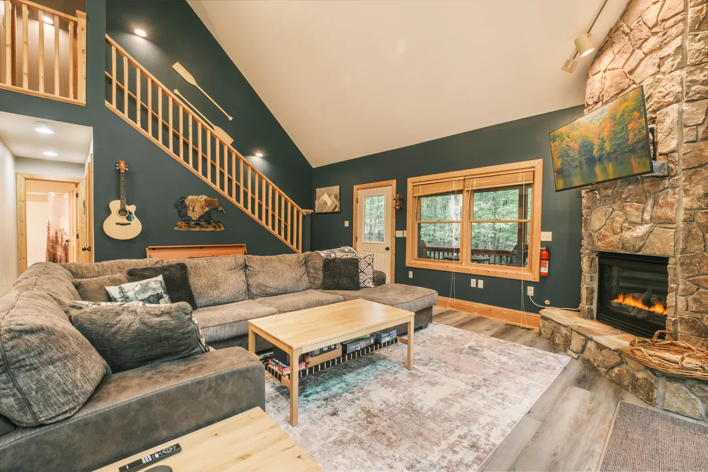 Living room with a large sectional sofa, wooden coffee table, stone fireplace, wall-mounted TV, and stairs with wooden railing.