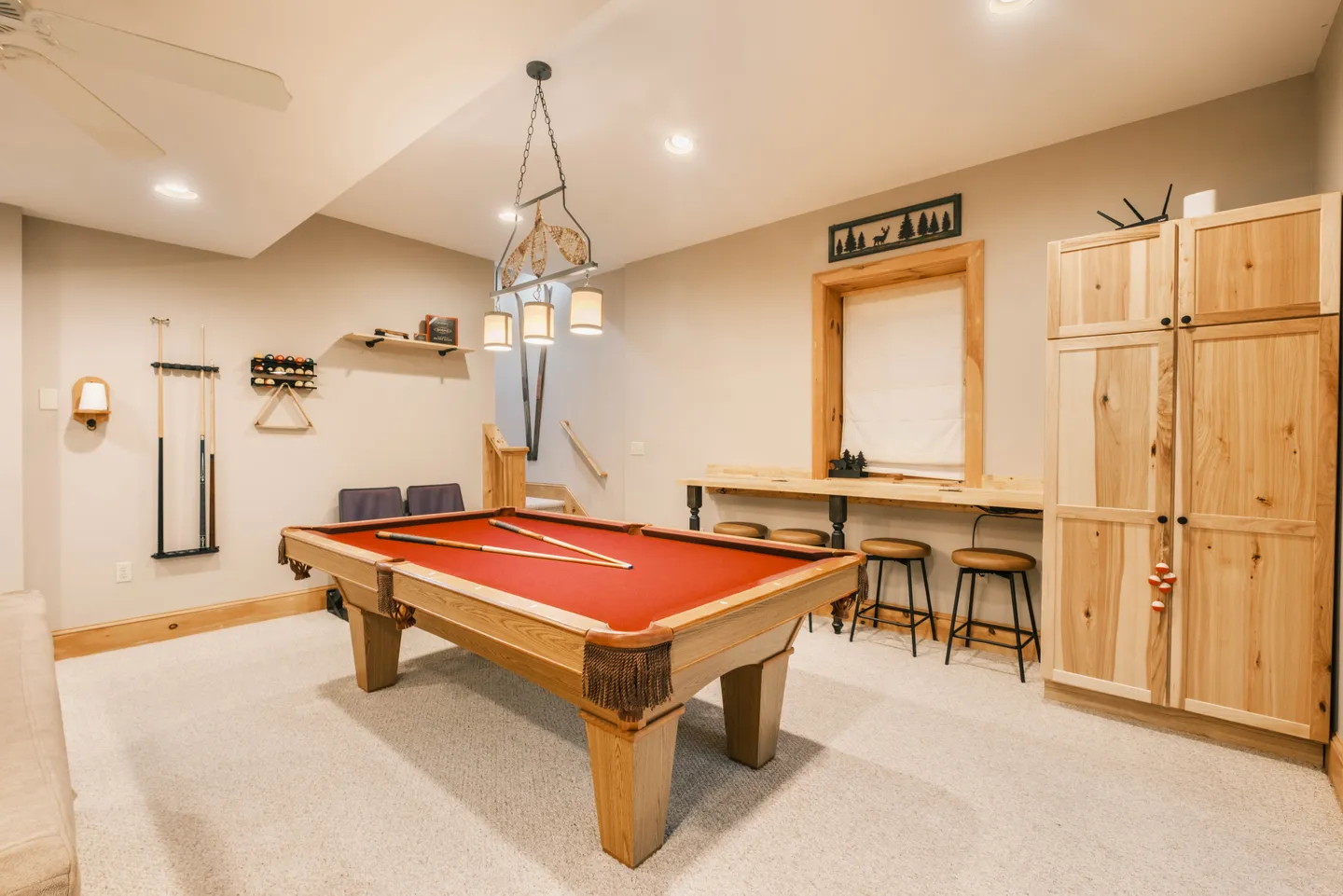 Game room featuring a wooden pool table with red felt, a wall-mounted cue rack, a wooden bar table with stools, and light wood storage cabinets.