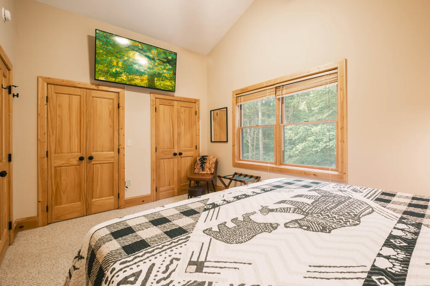Bedroom with patterned bear-themed quilt, mounted flat-screen TV, wood closet doors, and a window facing green trees.
