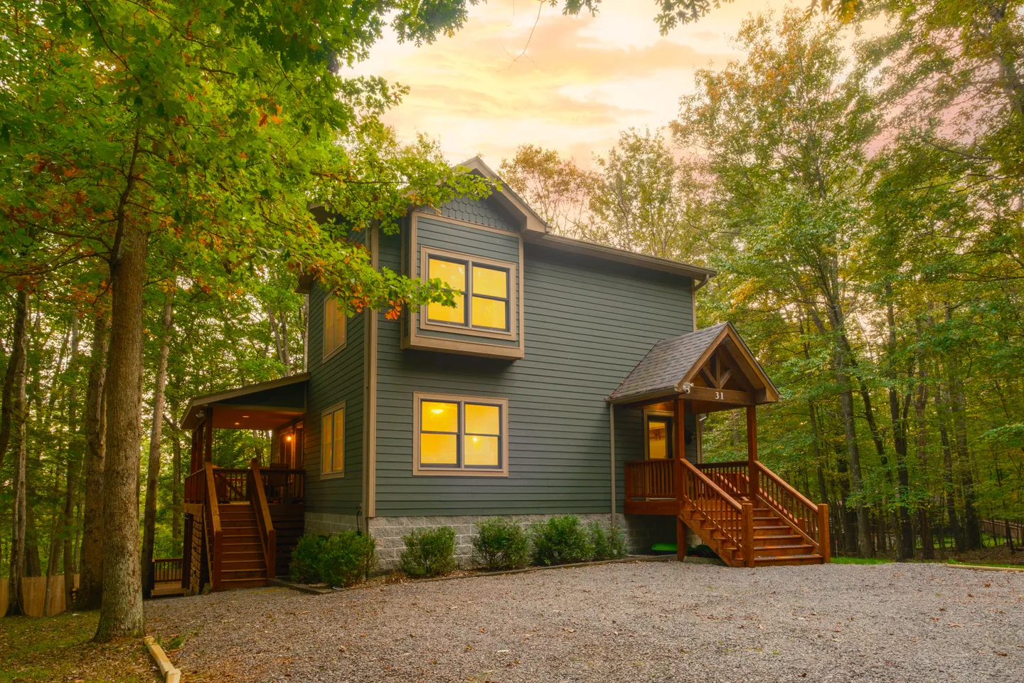 Exterior of a two-story dark green home with wood porch steps, gravel driveway, and surrounded by trees.