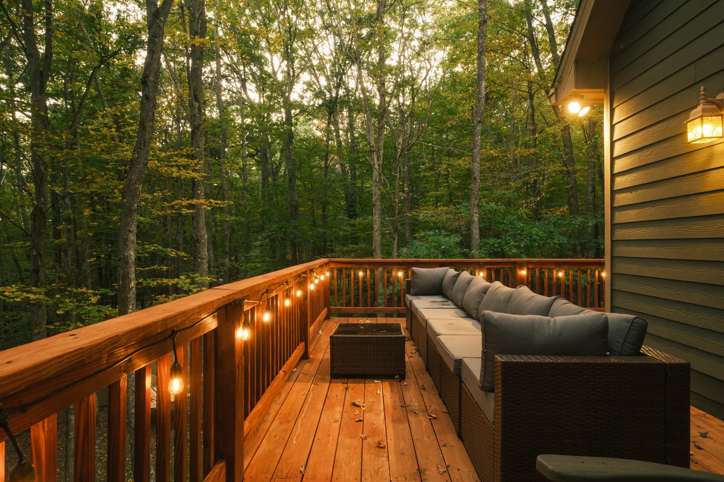 Outdoor wooden deck with a large wicker sectional sofa, string lights, and a view into dense green woods.