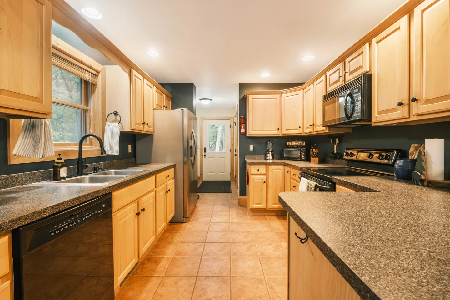 A kitchen with light wood cabinetry, speckled dark countertops, tile flooring, and standard appliances including a stainless steel refrigerator and black microwave.