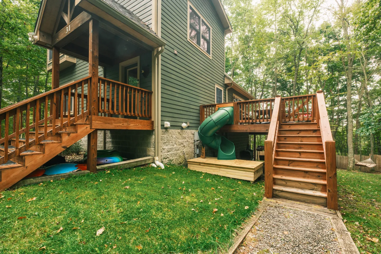 Rear exterior of a multi-level house with wooden decks, stairs, a green spiral slide, and a grass lawn.