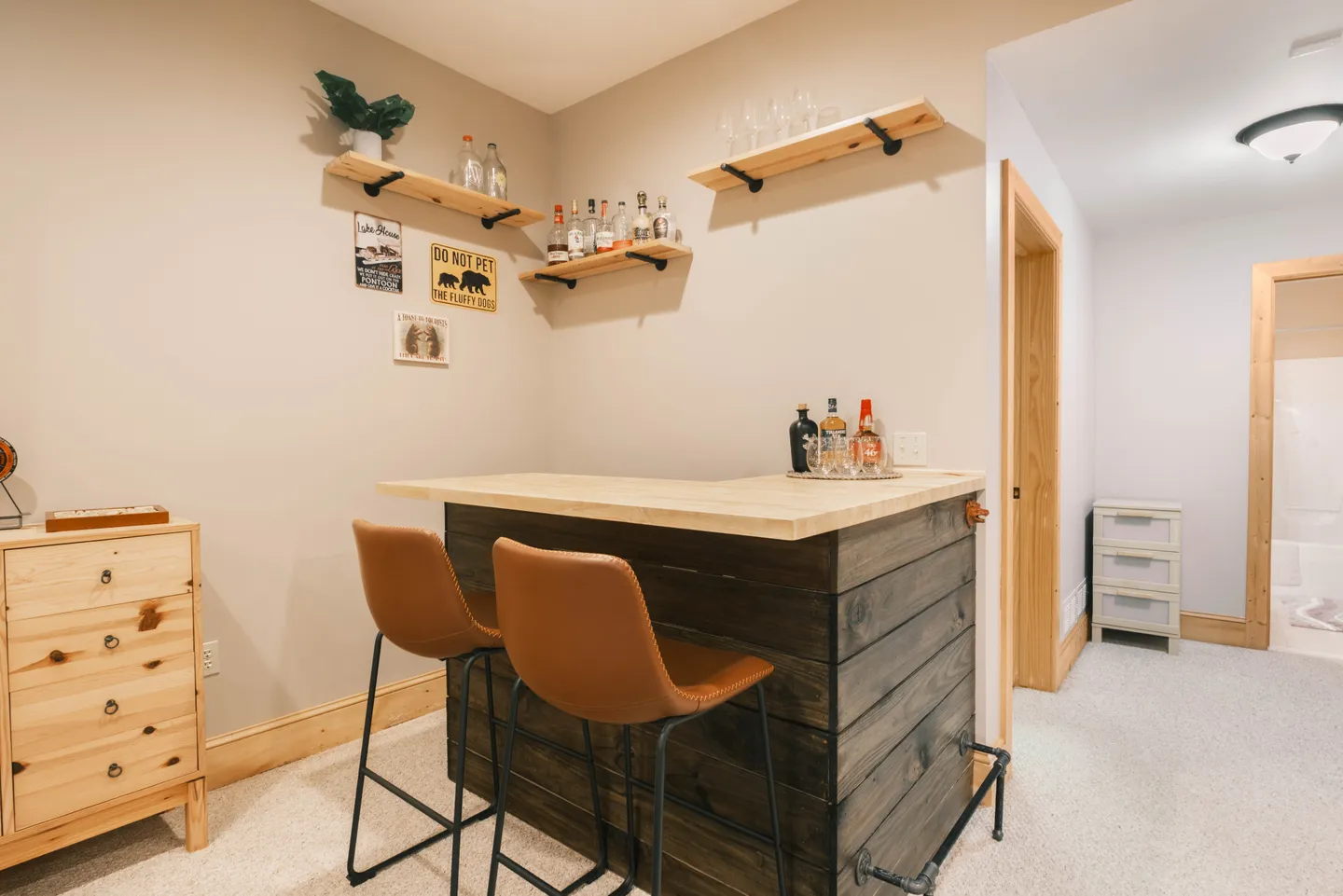A wooden home bar nook with two brown barstools, wood plank base, light wood countertop, and floating wall shelves.