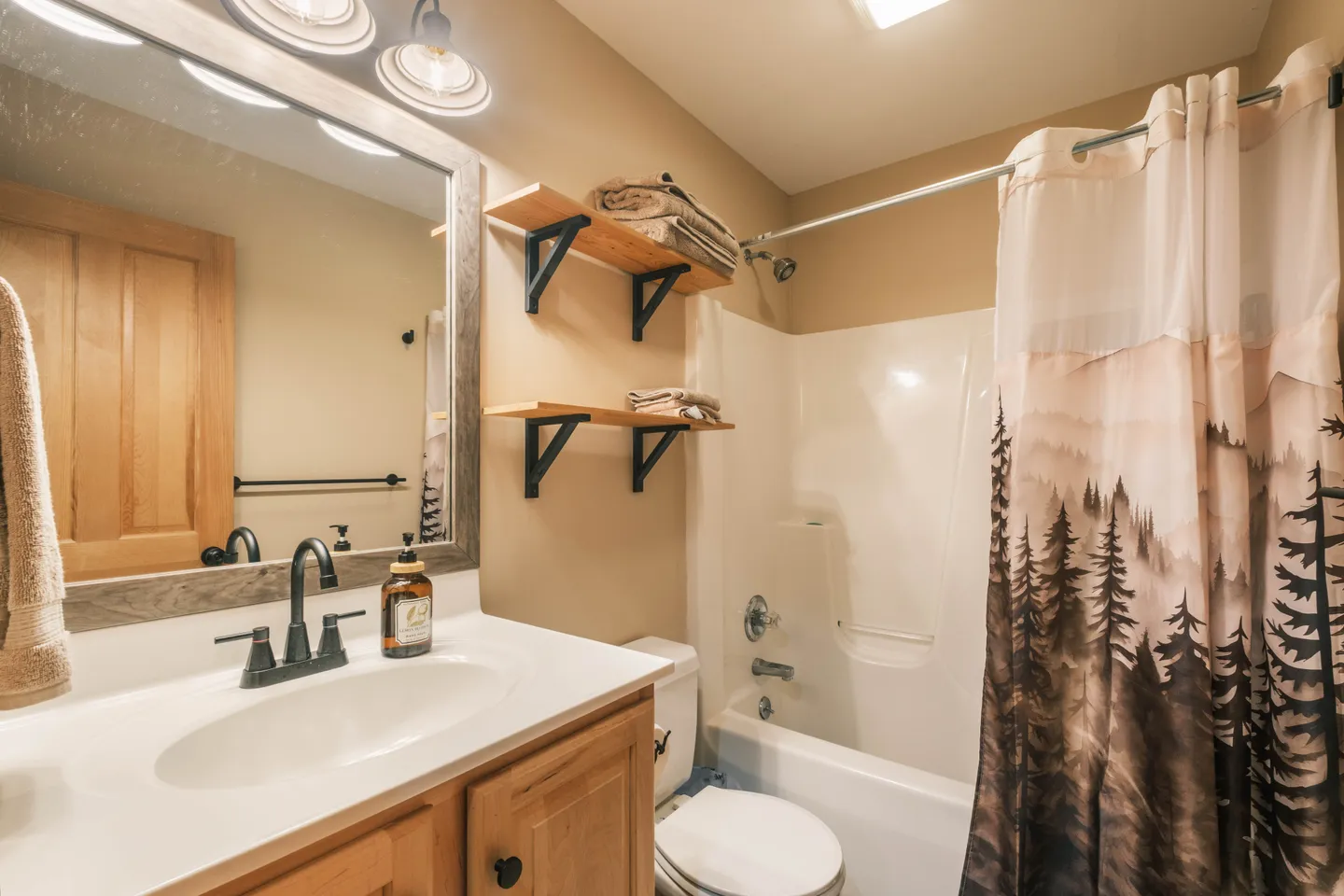 Bathroom with a wood vanity, framed mirror, toilet, open wall shelves, and a shower-tub combo with a pine tree shower curtain.