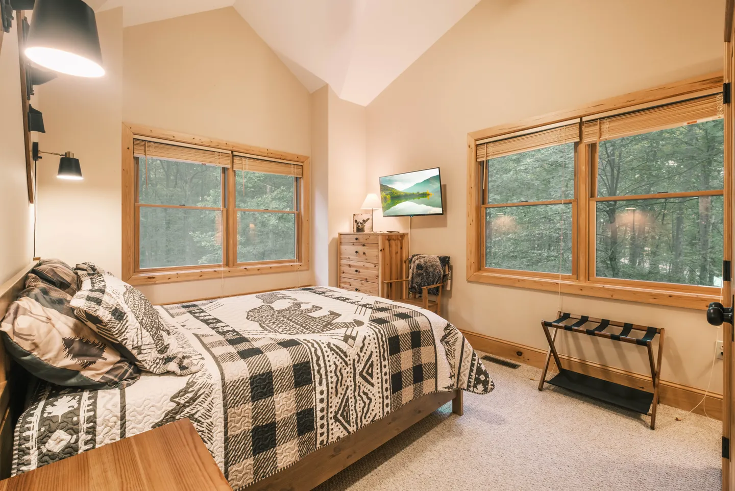 Bedroom with vaulted ceiling, large windows looking out to trees, and a bed with rustic patterned bedding.