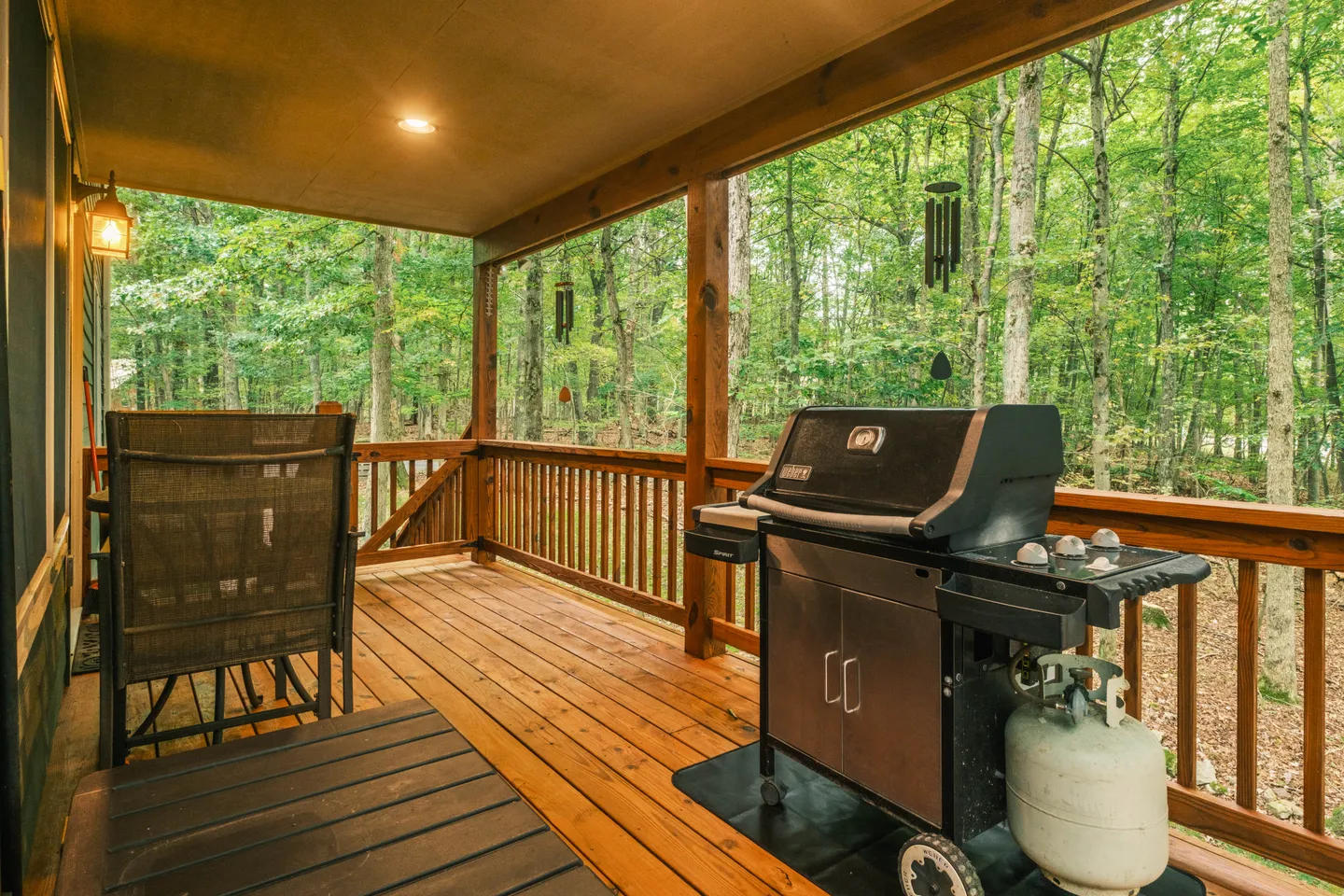 Covered wooden deck with a gas barbecue grill, seating, and a view of surrounding green forest.