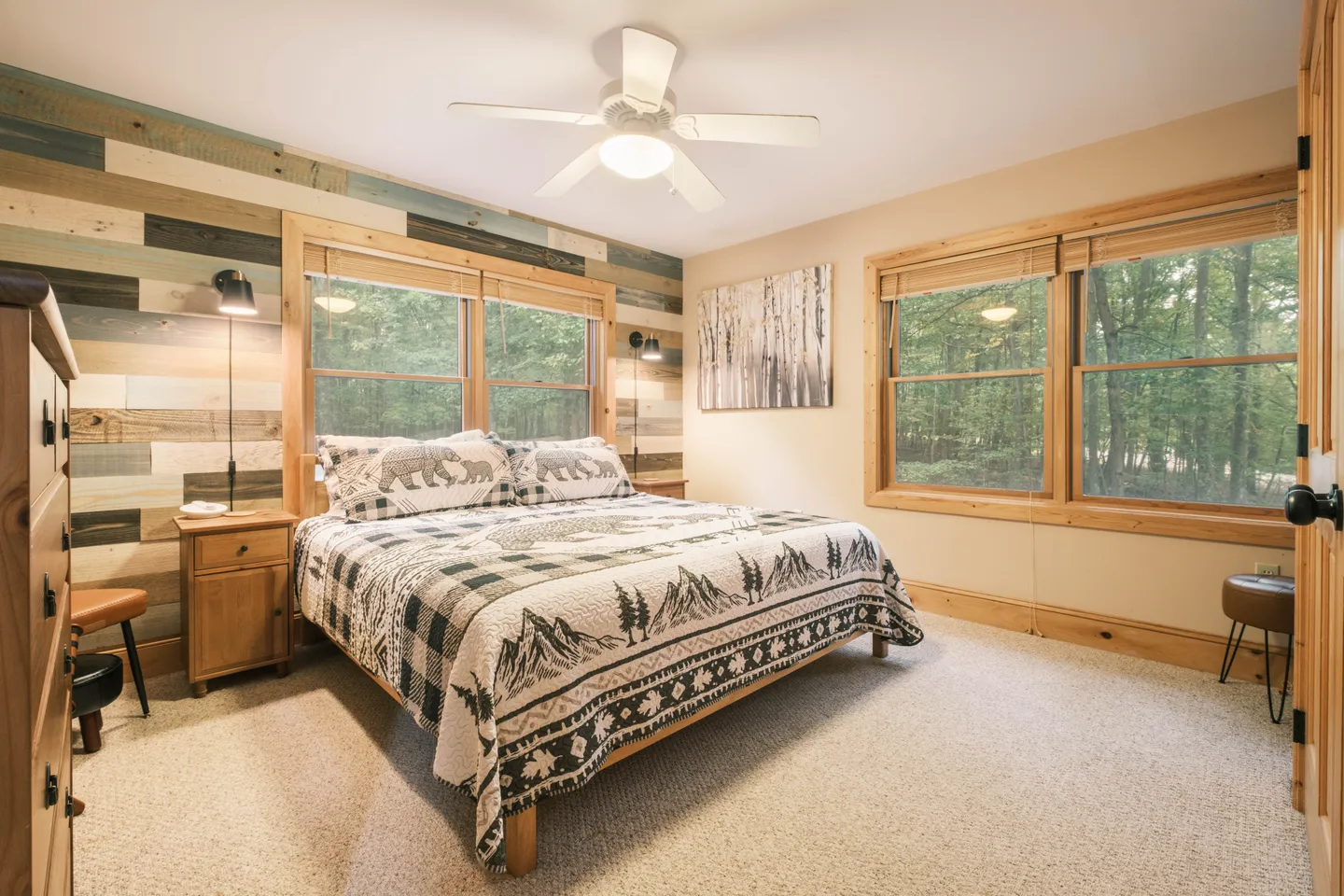 Bedroom with carpeted floors, rustic wood-plank accent wall, wood-trimmed windows with views of trees, and a bed with mountain-themed bedding.