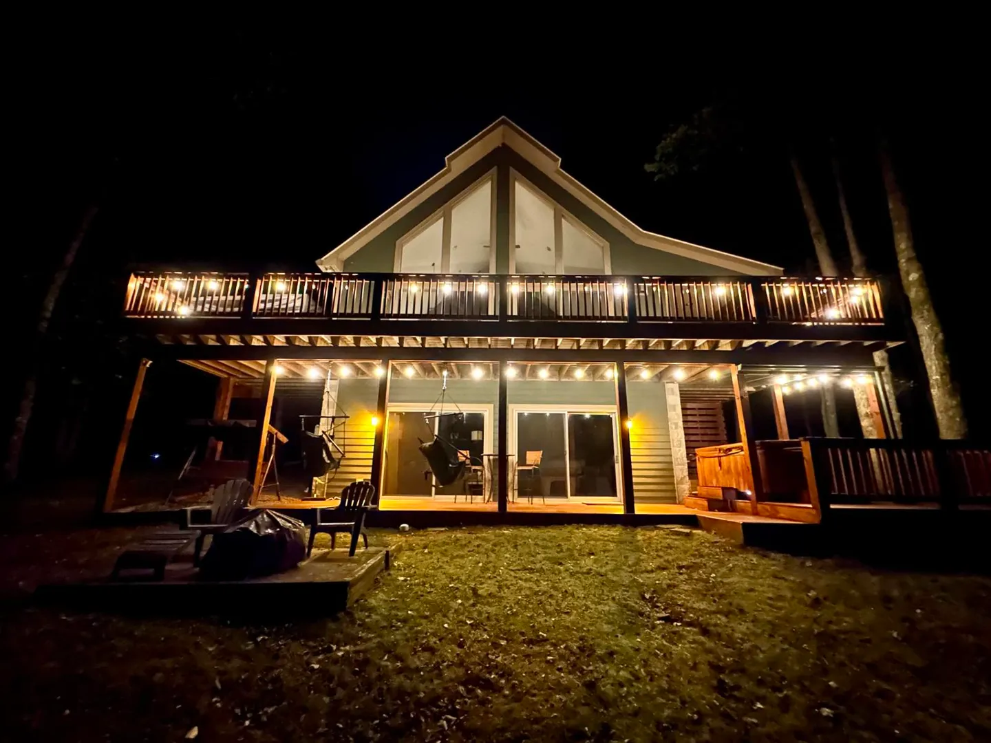 Night exterior view of a multi-level cabin with string lights on the upper deck and lower patio, hot tub, and yard.