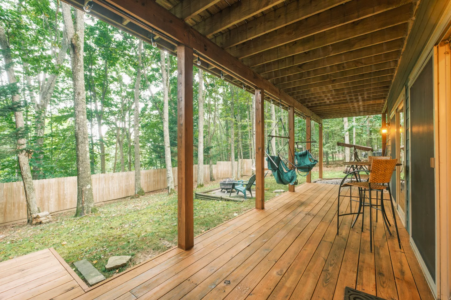 A covered wooden deck with hanging chairs overlooking a fenced-in wooded backyard.